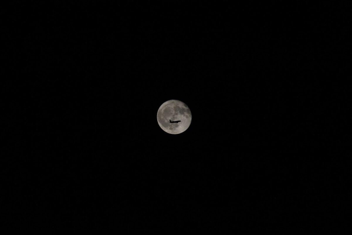 An airplane silhouette against a bright full moon in a clear night sky, showcasing travel and exploration.
